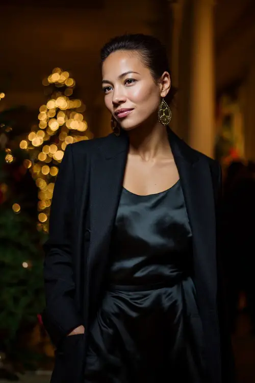 A woman wearing a sleek black satin dress with a structured long coat and bold earrings, standing at an upscale holiday event with warm ambient lighting