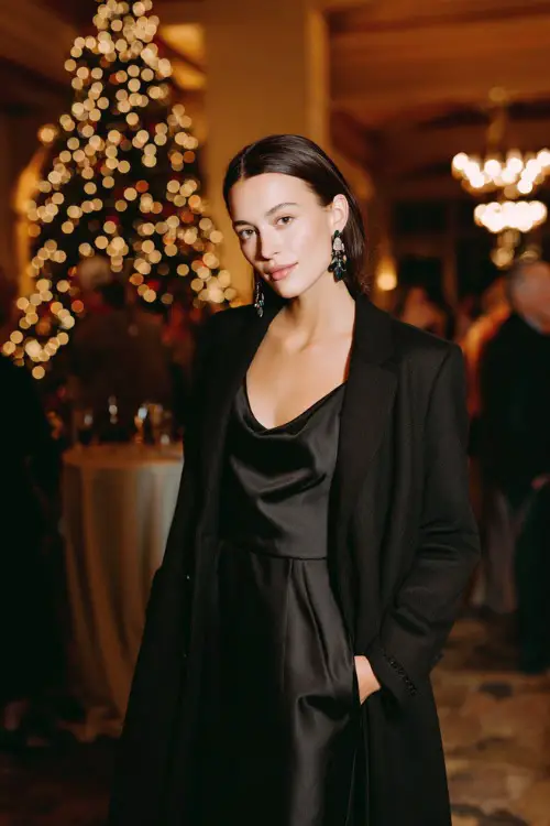 A woman wearing a sleek black satin dress with a structured long coat and bold earrings