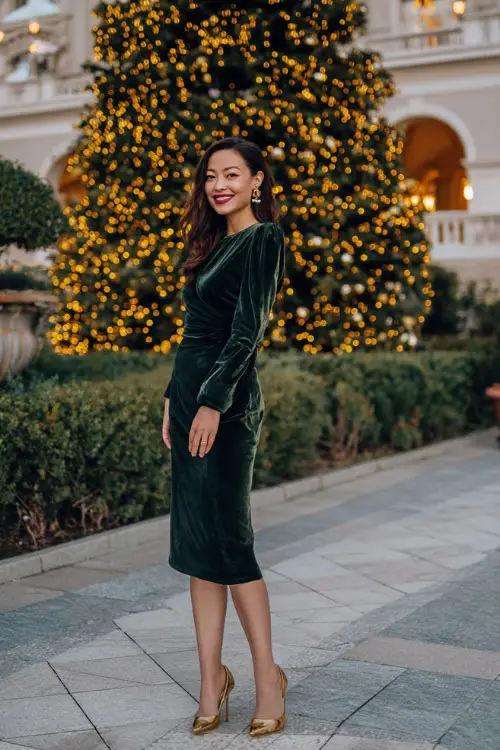 A woman wearing an emerald green velvet midi dress with long sleeves, gold heels, and pearl earrings, standing near a grand Christmas tree with warm golden lights