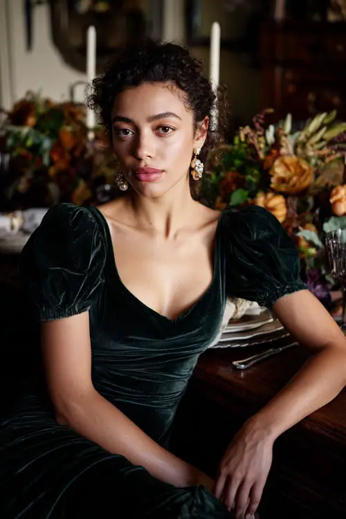 A woman wears a dark green velvet midi dress with puff sleeves and delicate earrings, sitting at a Thanksgiving dinner table with floral centerpieces