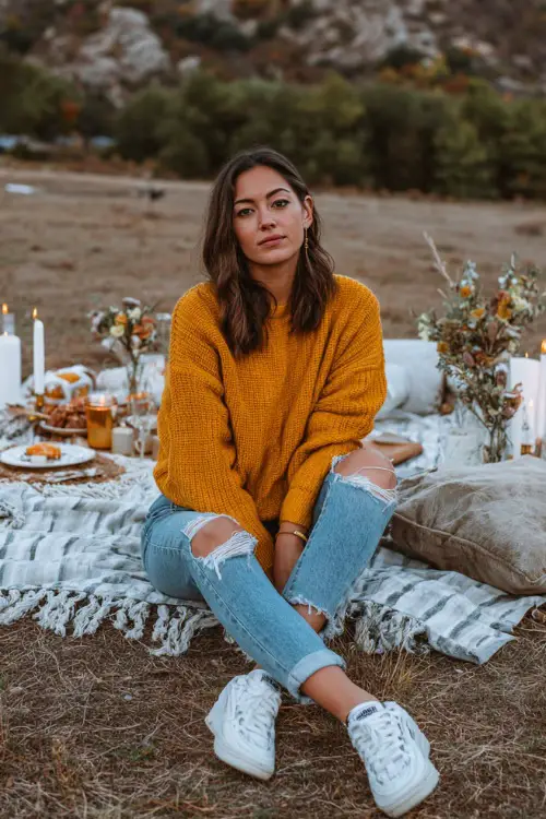 A woman wears a mustard sweater with ripped jeans and white sneakers