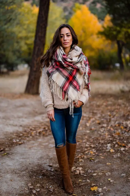 A woman wears a plaid blanket scarf over a chunky beige sweater, blue jeans, and tall brown boots, standing outdoors in a park filled with fall colors 