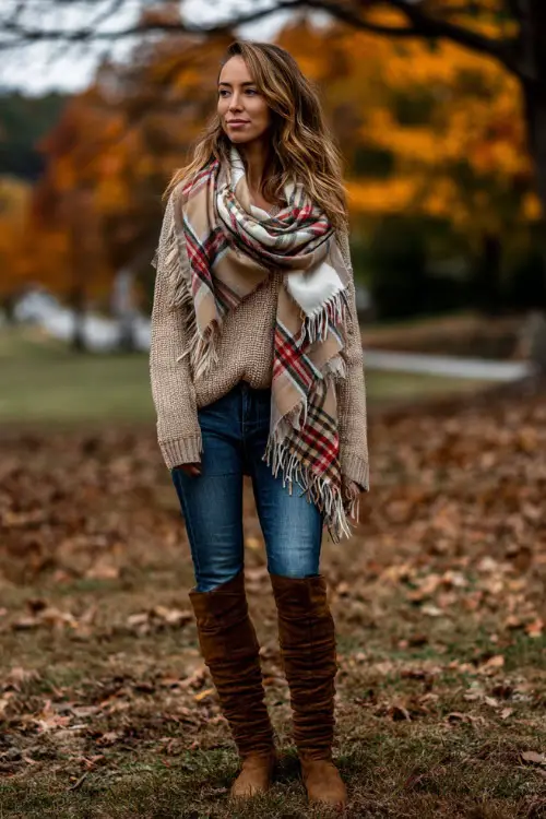A woman wears a plaid blanket scarf over a chunky beige sweater, blue jeans, and tall brown boots, standing outdoors in a park filled with fall colors
