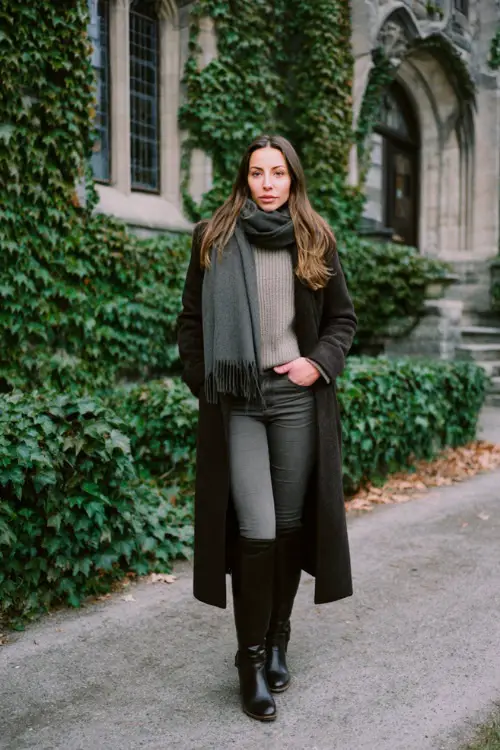 A woman wearing straight-leg jeans styled with a long wool coat, fitted knit sweater, leather riding boots, and a silk scarf, standing near an ivy-covered building in winter