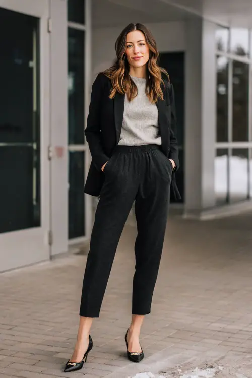 A full-body image of a midsize woman dressed in a monochrome business casual outfit featuring a blazer and matching trousers, styled with a lightweight knit top and pointed flats