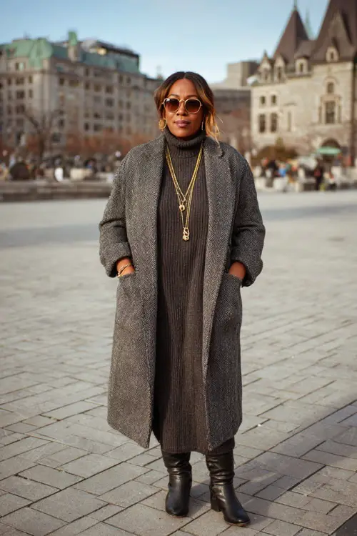 A plus-size woman wearing a long wool coat layered over a ribbed knit dress, styled with leather ankle boots and minimal gold jewelry, standing in a quiet winter square