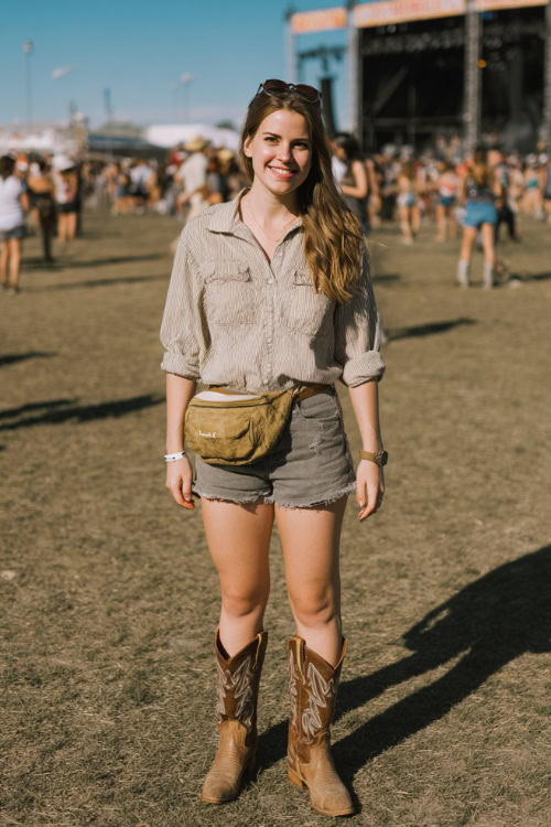 A woman in her 30s wears a relaxed spring country concert outfit with high-waisted denim shorts, a lightweight button-down shirt, classic cowboy boots