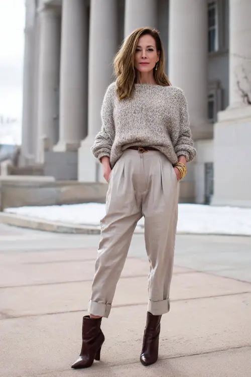 A woman in her 40s wears a sophisticated boho outfit featuring a textured knit sweater tucked into tailored trousers, heeled ankle boots, and elegant accessories