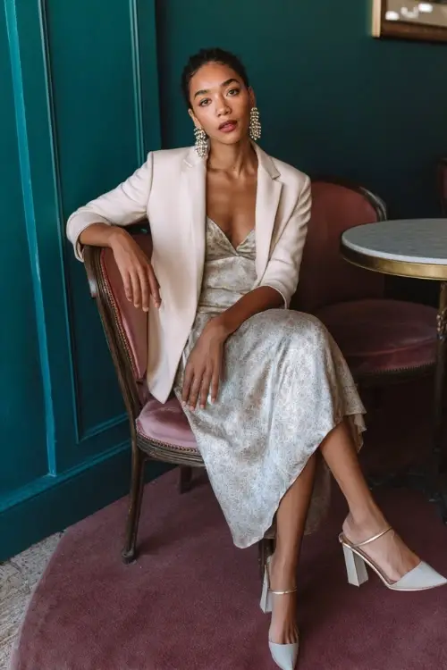 A woman over 30 wears a chic formal boho ensemble with a silk midi dress, a soft tailored blazer, block heels, and statement earrings, styled in a classy indoor event venue