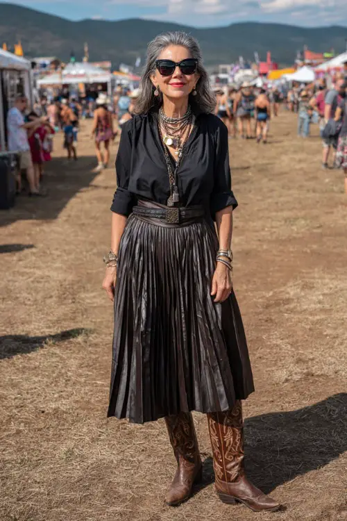 A woman over 40 wears a polished country concert outfit with a pleated midi skirt paired with a fitted blouse, tall cowboy boots, and layered necklaces