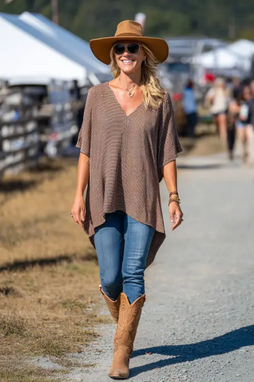 A woman over 50 wears a relaxed country concert outfit with a flowy tunic top paired with straight-leg denim jeans, tan cowboy boots, and a wide-brim felt hat 