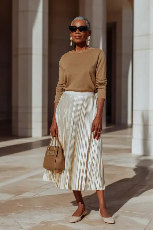 A woman over 50 wears an elegant boho ensemble with a pleated midi skirt, a fitted knit top, low block heels, and statement earrings 