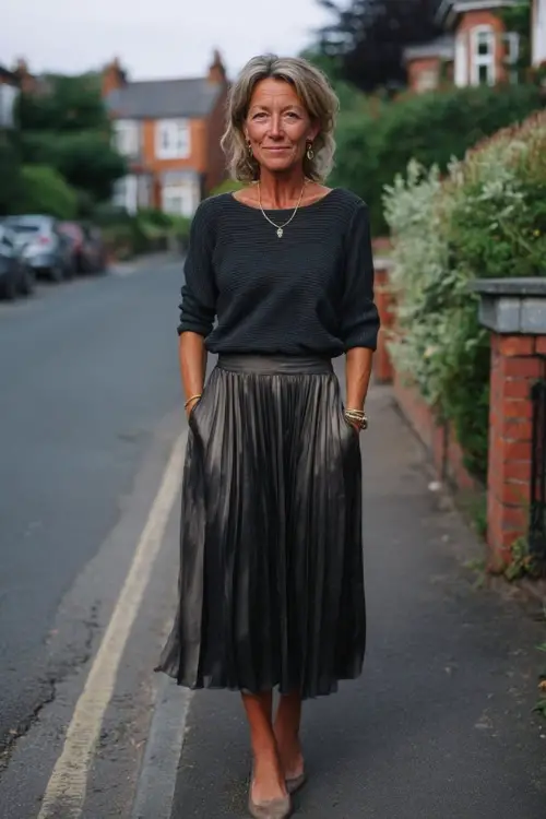 A woman over 50 wears an elegant boho outfit combining a pleated midi skirt, a comfortable knit top, low block heels, and subtle statement earrings, styled in a stylish residential street