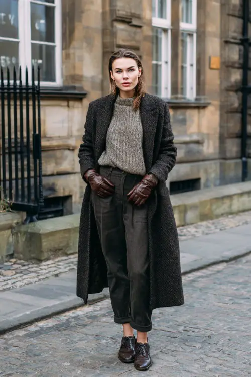 A woman wearing a long wool coat layered over a cashmere sweater and straight-leg jeans, styled with leather loafers and leather gloves, standing near historic buildings in winter