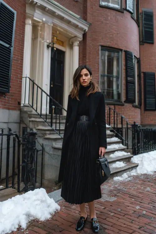 A woman wearing a pleated midi dress styled with a structured blazer, fine knit layer, leather loafers, and a top-handle handbag, standing near classic townhouses in winter