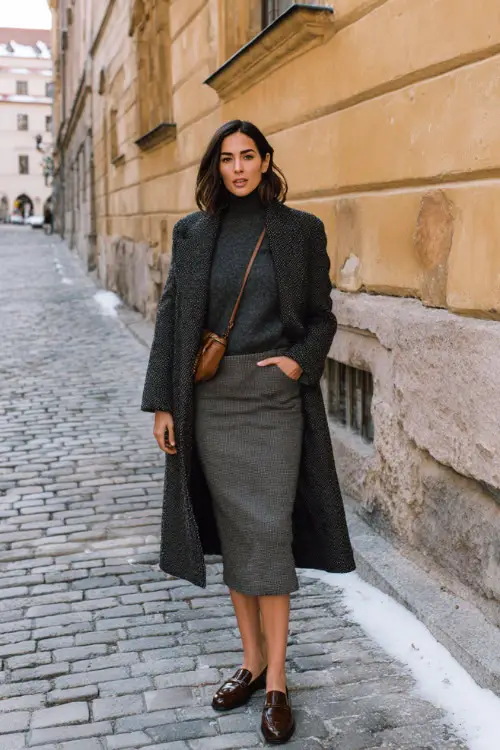 A woman wearing a wool midi skirt styled with a fitted knit sweater, casual wool coat, leather loafers, and a simple crossbody bag, standing along a cobblestone street in winter