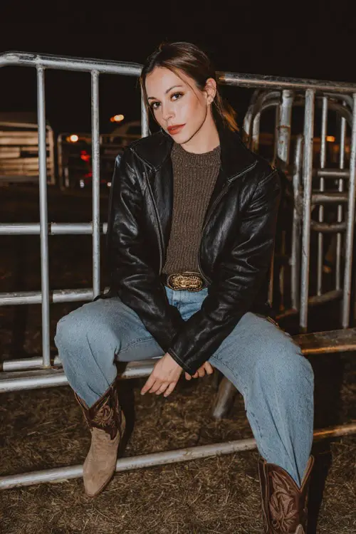 A woman wears a chic winter country concert outfit with a leather jacket over a thick knit sweater, straight jeans, western boots, and statement belt 