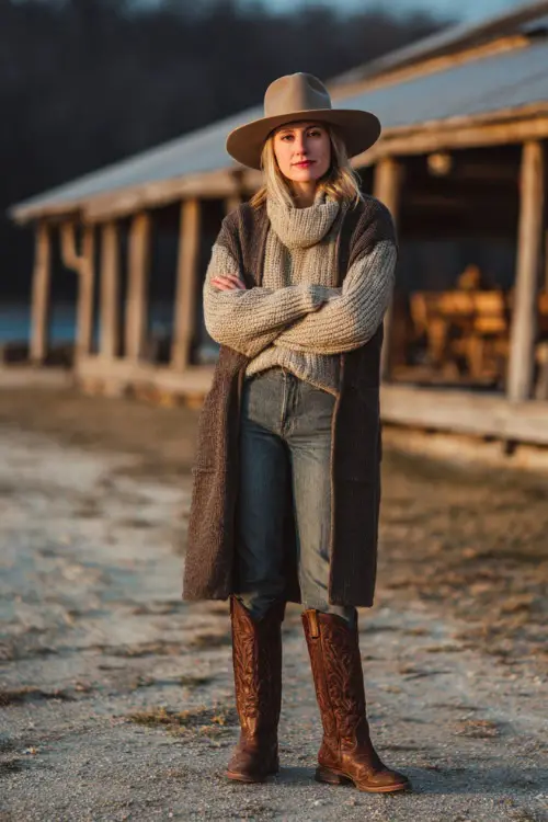 A woman wears a cozy country concert outfit with a chunky knit sweater, straight-leg jeans, tall brown cowboy boots, and a wool felt hat, layered with a long tailored coat 