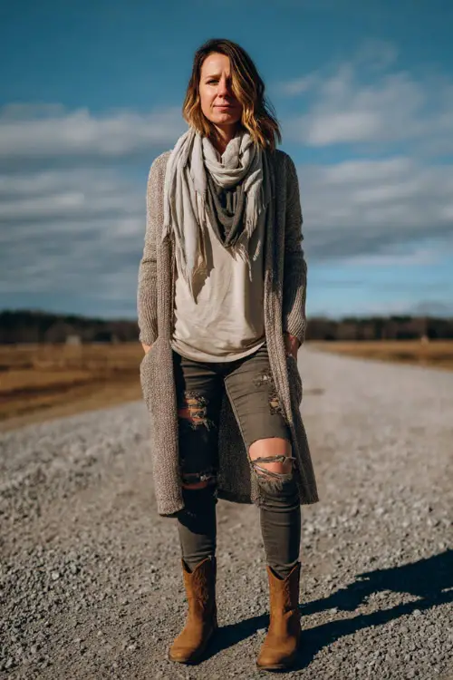 A woman wears a cozy layered country concert outfit featuring a long cardigan, fitted thermal top, ripped jeans, ankle cowboy boots, and a scarf