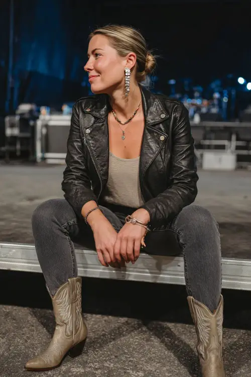 A woman wears a modern country concert look with a leather jacket over a neutral top, dark-wash jeans, pointed western boots, and statement earrings