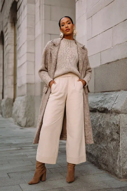 A woman wears an elegant boho winter outfit combining a textured knit sweater, wide-leg pants, a belted coat, and block-heel boots