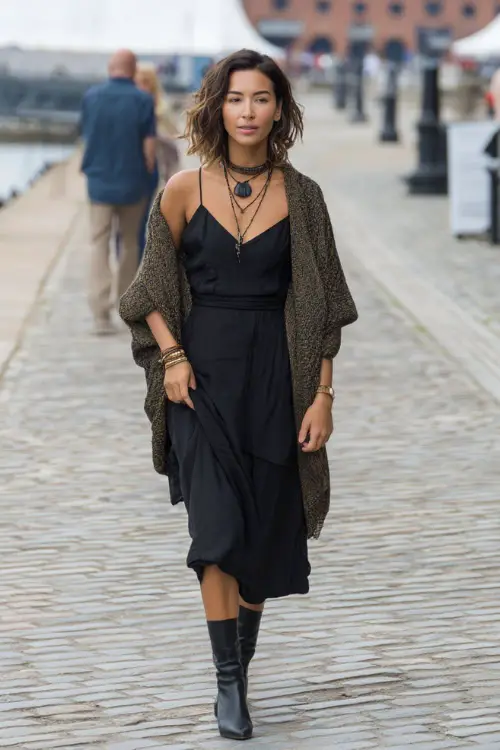 A woman wears an elevated chic boho outfit with a monochrome midi dress, a soft shawl-style jacket, ankle boots, and layered jewelry, styled in a refined urban walkway
