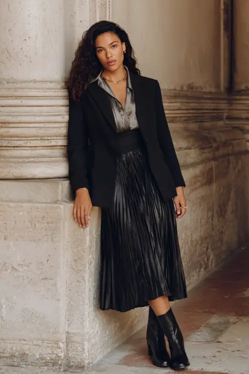 A curvy woman wears a pleated midi skirt with a silk blouse and structured blazer styled with Chelsea boots, posing near historic architecture, full-body editorial fashion shot