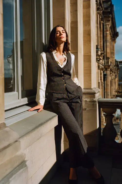 A woman over 30 wears a pinstripe vest layered over a silk blouse with tailored trousers and low heels, photographed on a city balcony overlooking classic architecture