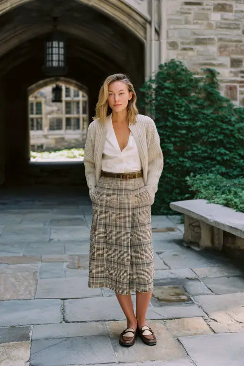 A woman over 30 wears a plaid midi skirt with a lightweight cardigan draped over shoulders and loafers, photographed in a university courtyard, full-body fashion capture 