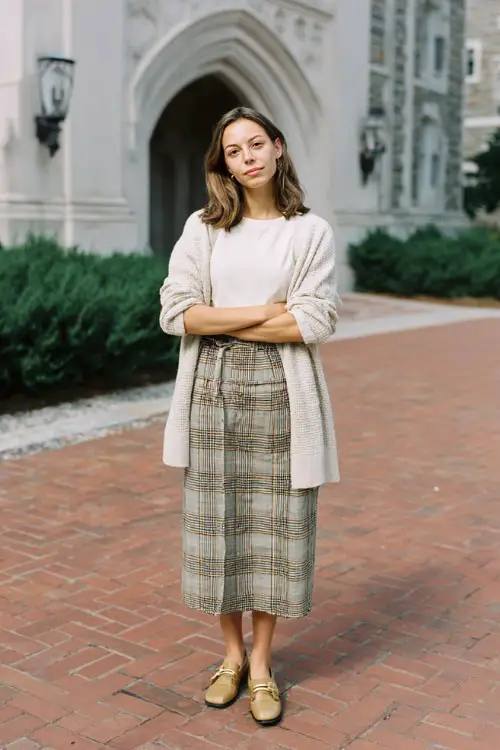 A woman over 30 wears a plaid midi skirt with a lightweight cardigan draped over shoulders and loafers, photographed in a university courtyard, full-body fashion capture