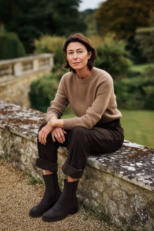 A woman over 40 wears a cashmere sweater with tailored trousers and suede Chelsea boots, photographed in a countryside estate garden, full-body portrait 