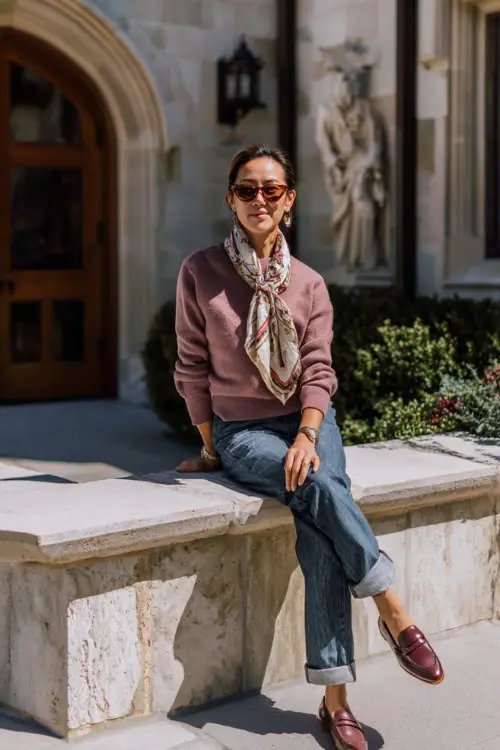 A woman over 40 wears a fine knit sweater with a silk scarf, straight jeans, loafers, styled in a university courtyard, full-body fashion photography, natural soft light