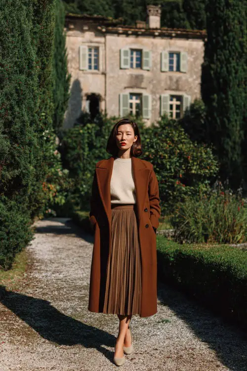 A woman over 40 wears a long wool coat layered over a pleated midi skirt, cashmere sweater, and low heels, photographed in a countryside estate garden 