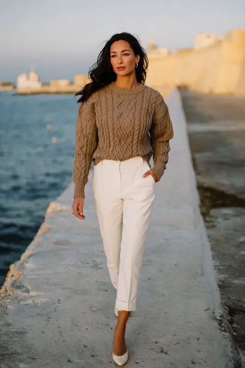 A woman wears a classic cable-knit sweater with white trousers and ballet flats, walking along a coastal promenade, full-body lifestyle fashion photography