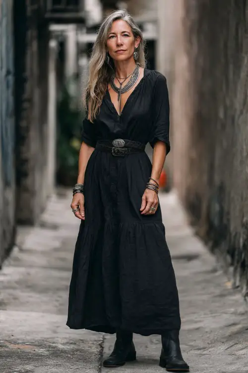 A woman over 40 wears a black flowy boho maxi dress paired with a leather belt and ankle boots, styled with layered silver jewelry
