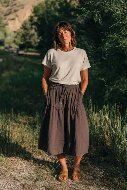 A woman over 40 wears a casual tiered midi skirt with a relaxed t-shirt tucked in, paired with slip-on sandals and minimal jewelry, standing in a sunlit park with greenery