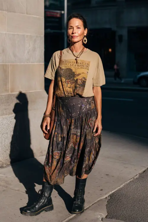A woman over 40 wears a loose graphic tee tucked into a dark boho midi skirt with subtle prints, paired with chunky boots and statement earrings