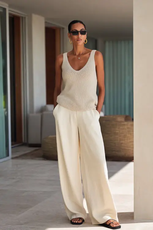 A woman over 40 wears a relaxed knit tank top with flowy cream wide-leg pants, styled with slide sandals and oversized sunglasses, standing in a modern outdoor patio with soft neutral tones