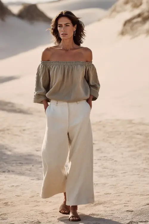 A woman over 40 wears a relaxed off-shoulder linen top with wide-leg pants in soft neutral shades, styled with minimal accessories and flat sandals