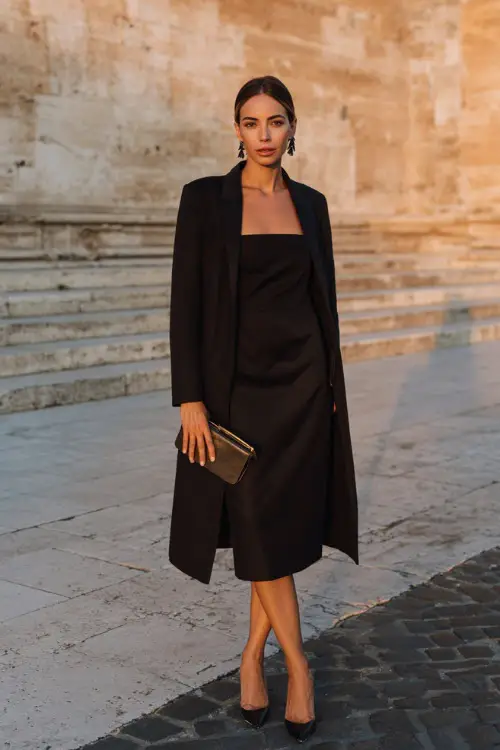A woman wears a black midi dress layered with a tailored coat, styled with pointed-toe heels and a sleek leather clutch, accessorized with statement earrings