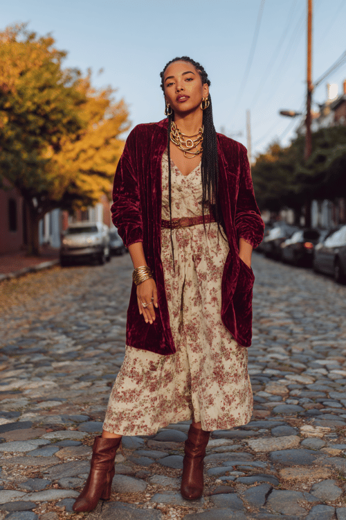 Black woman with long braided hair wearing a rich burgundy velvet kimono jacket over a cream floral midi dress, stacked gold rings and layered chain necklaces