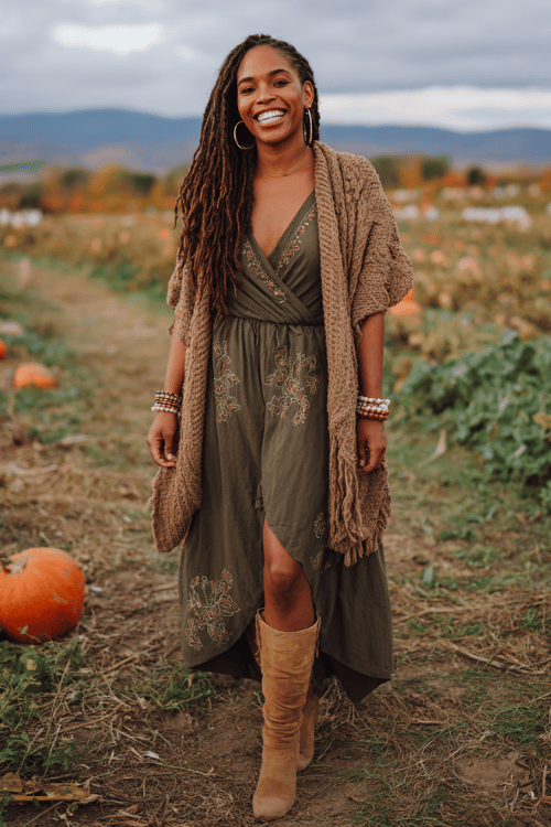 Black woman with long loc hairstyle wearing a deep olive wrap maxi dress with floral embroidery and flutter sleeves, a chunky knit earth-toned cardigan draped over her shoulders