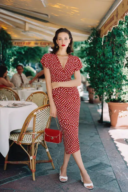 Full body editorial fashion photograph of a poised woman wearing a timeless red and white polka dot wrap midi dress with cap sleeves