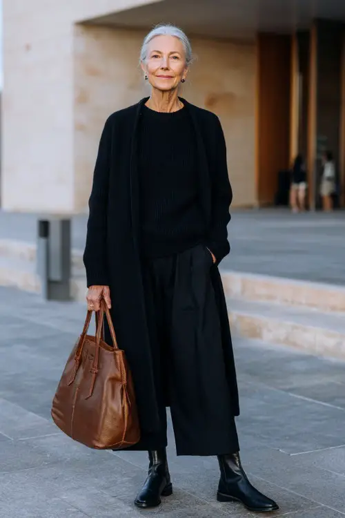 Full body editorial photograph of a gracefully sophisticated woman over 50 dressed in a classic monochrome all-black ensemble featuring a relaxed cashmere crewneck sweater 