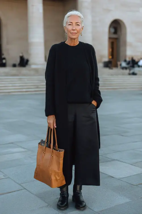 Full body editorial photograph of a gracefully sophisticated woman over 50 dressed in a classic monochrome all-black ensemble featuring a relaxed cashmere crewneck sweater