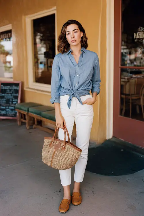 Full body editorial shot of a laid-back elegant woman wearing a classic light blue chambray button-down shirt tied at the waist over a pair of well-fitted white slim ankle-length trousers 