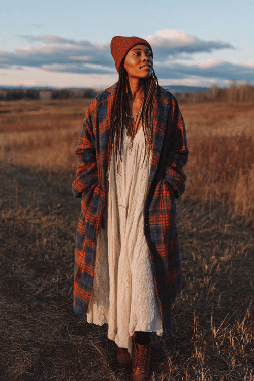 a Black woman with long highlighted locs wearing a cozy oversized plaid blanket coat in rust and navy over a long cream lace maxi dress, tall chocolate brown lace-up leather boots