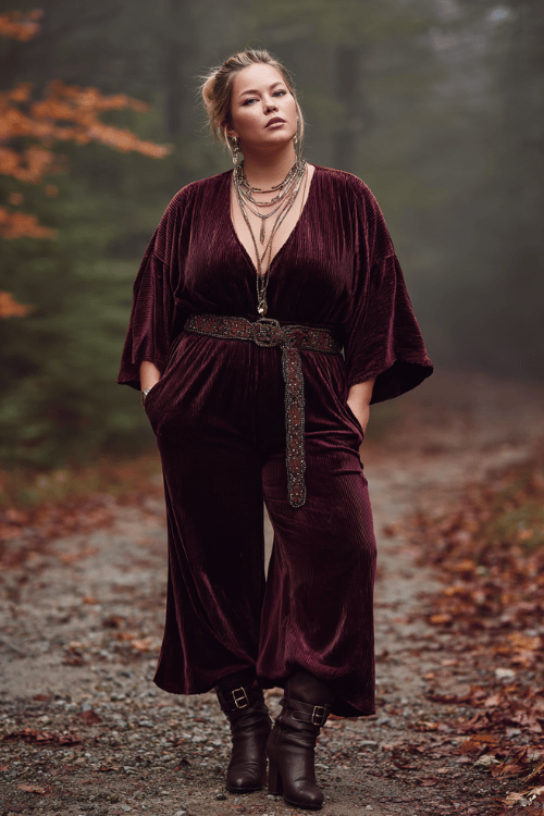 a plus-size woman in a cozy dark plum velvet flared jumpsuit with wide sleeves, a braided leather belt, layered crystal necklaces, and ankle boots with buckle detail