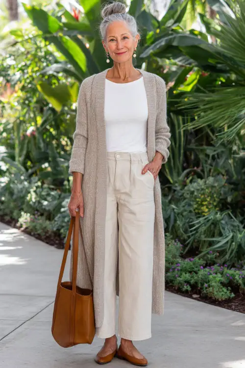 a poised woman over 50 wearing a classic long open-front soft oatmeal cardigan layered over a fitted white linen t-shirt and high-waisted straight-leg light beige chino trousers