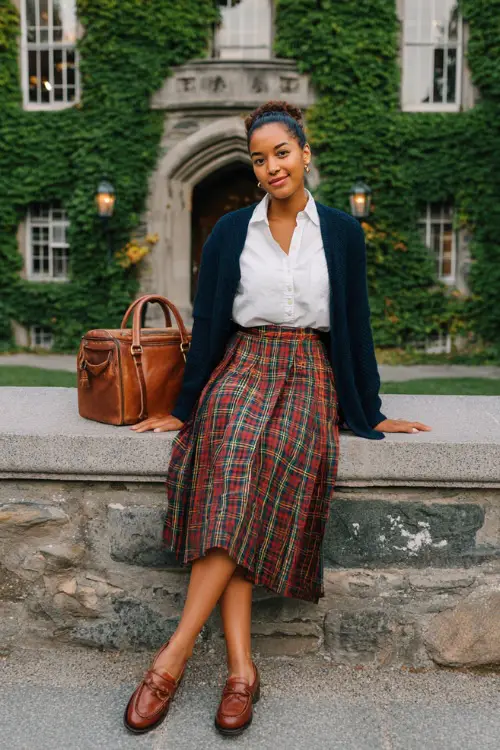 a stylish woman wearing a classic high-waisted pleated tartan plaid midi skirt in warm red and green tones paired with a fitted crisp white button-down shirt tucked in